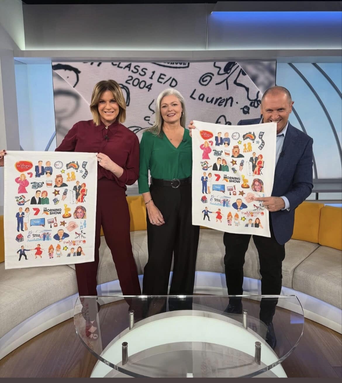 Three people stand in a studio, smiling and holding up illustrated posters depicting various cartoon figures and logos.