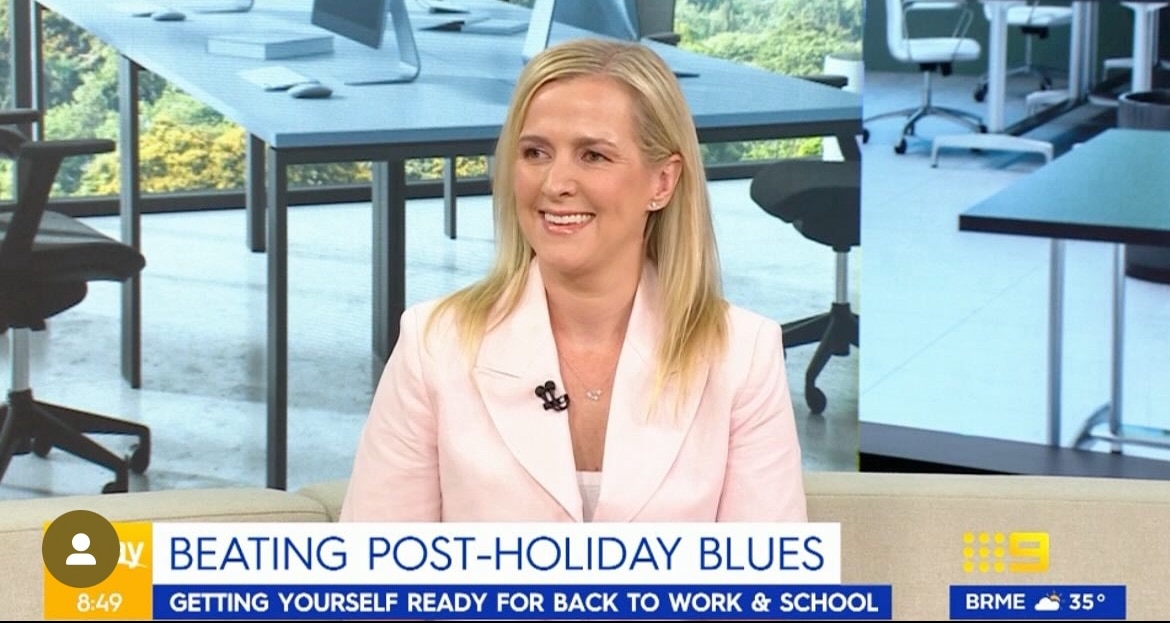 A woman in a light pink blazer sits and smiles during a morning TV segment about beating post-holiday blues, with an office setting shown in the background.